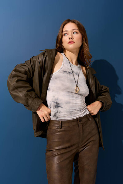 Красивая молодая женщина уверенно позирует в коричневой кожаной одежде на синем фоне.