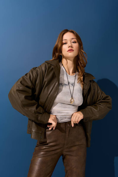 Красивая молодая женщина уверенно стоит в стильной коричневой кожаной одежде на синем фоне.