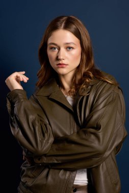 Genç bir kadın açık mavi bir arka plana karşı kahverengi deri bir elbise modelliyor..