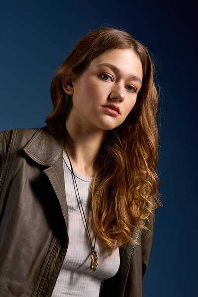 Уверенная молодая женщина в коричневой кожаной одежде позирует в синей студии, излучая осенние вибрации.