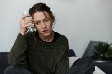 Genç bir kadın evdeki kanepede oturur, elinde bir bardak su, derin düşüncelere dalmış ve üzgün hisseder..