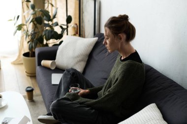 Genç bir kadın kanepesinde yalnız başına oturuyor, düşüncelere dalmış, elinde bir bardak su tutuyor..