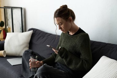 Genç bir kadın koltuğunda sıkıntılı bir şekilde oturuyor, telefonunu kullanıyor ve elinde bir bardak su tutuyor..