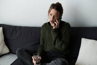 Genç bir kadın kanepesinde oturuyor, elinde bir bardak su tutuyor ve bir telefon görüşmesi sırasında üzüntüsünü dile getiriyor..