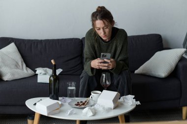 Bir kadın kanepeye oturur, atılmış mendiller ve yiyecek kaplarıyla çevrili, düşüncelere dalmış..