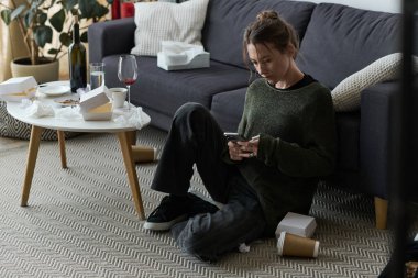 Genç bir kadın yerde oturuyor, boş konteynırlar ve mendillerin arasında telefonunu kaydırıyor..