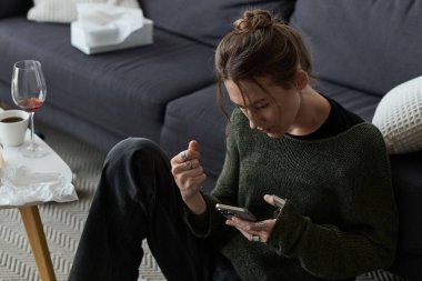 Genç bir kadın kanepede oturur, evde tek başına telefonunu kontrol ederken kendini kötü hisseder..
