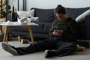 Oturma odasının zemininde oturan genç bir kadın üzgün bir şekilde telefonuna sarılmış..