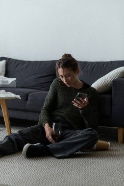 Genç bir kadın elinde şarapla yerde oturur loş bir oturma odasında telefonuna bakar..