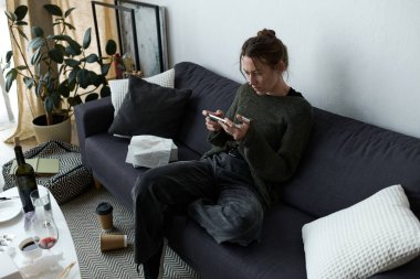 Genç bir kadın kanepesinde oturuyor, telefonuna dalmış, yalnızlıkla savaşıyor..