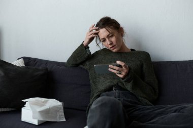 Kanepede oturan genç bir kadın, telefonuna bakarken düşüncelere dalmış, bunalmış hissediyordu..