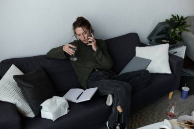 Genç bir kadın kanepesinde oturuyor, elinde bir bardak su tutuyor ve telefonda konuşuyor..