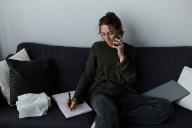 Genç bir kadın koltukta oturur, telefonda konuşurken düşüncelere dalmış ve notlar alır..