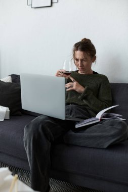 Genç bir kadın dizüstü bilgisayarını kullanırken ve elinde içki tutarken üzgün bir şekilde koltuğunda oturuyor..