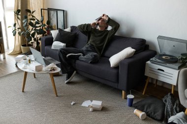 Genç bir kadın, karanlık bir kanepede oturur. Dağınık bir yaşam alanında üzüntü belirtileri gösterir..