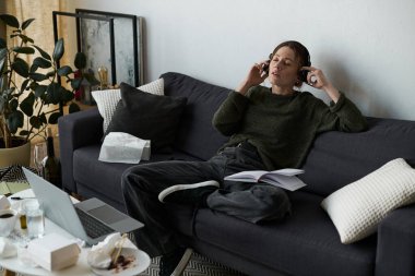 Genç bir kadın kanepesinde dinleniyor, kulaklık takıyor ve evdeki düşüncelerinde kayboluyor..