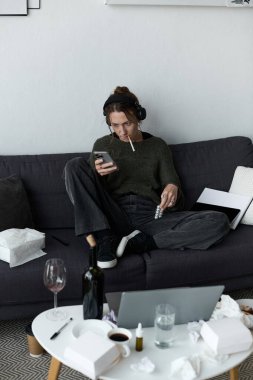 Kanepede oturan genç bir kadın kendini yorgun hisseder ve boş mendillerle çevrili telefonunu kullanır..