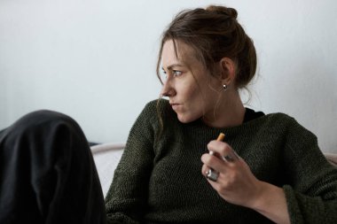 Genç bir kadın evde tek başına oturuyor, elinde sigara tutarken üzüntüsünü yansıtıyor..