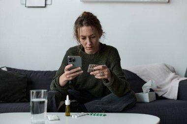 Genç bir kadın evdeki kanepede oturur, telefonuna kasvetli bir ifadeyle bakar, elinde haplarla..