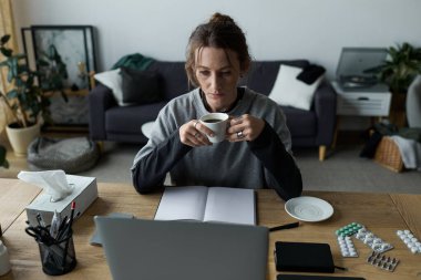 Rahat bir oturma odasında, genç bir kadın masasında tek başına kahvesini yudumlarken dalgın görünüyor..