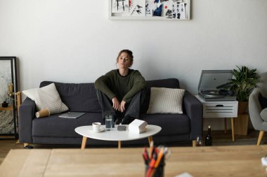 Oturma odasındaki kanepede oturan genç bir kadın. Etrafı sessiz ve yansıtıcı bir atmosferle çevrili..