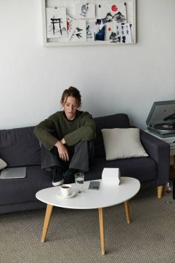 Genç bir kadın bir kanepede sessizce oturur, düşüncelere dalmış, sakinleştiren bir ev ortamıyla çevrili..