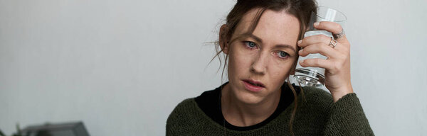 A young woman sits alone at home, looking pensive and weary, holding a glass of water to her head.