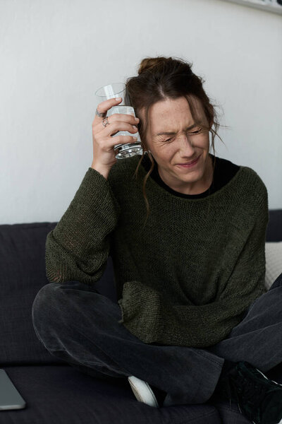 In a cozy living room, a young woman sits distressed, clutching a glass of water.