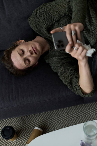 A young woman lies on her couch, absorbed in her phone, embodying feelings of sadness and isolation.