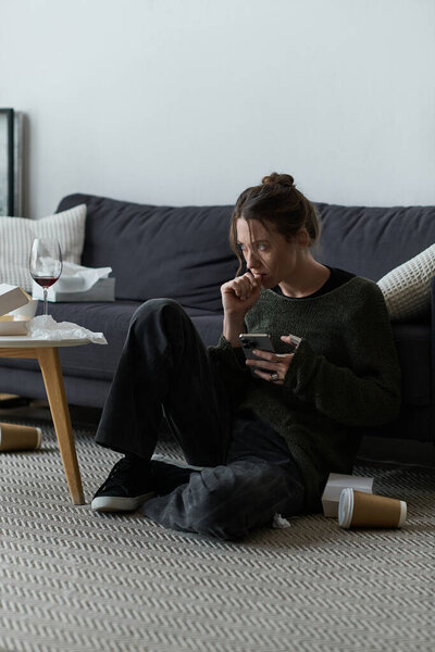 In a simple living room, a young woman sits on the floor, looking somber while using her phone.