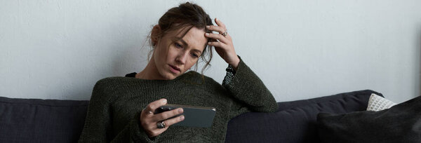 A young woman sits on a couch, deep in thought, checking her phone with a sad expression.