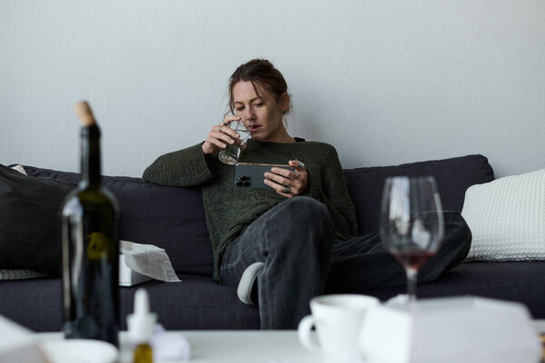 Young woman sitting on a couch, sipping water and staring at her phone, feeling alone.