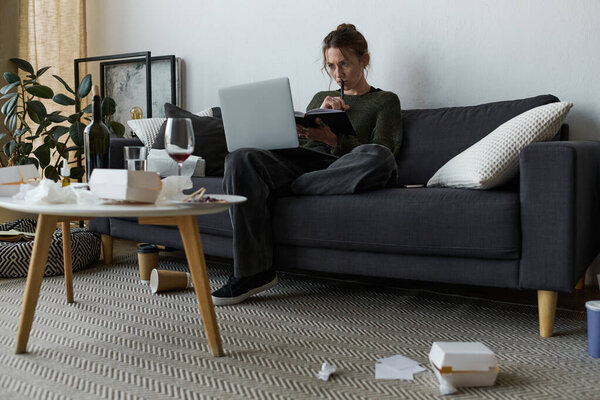 В уютной гостиной молодая женщина сидит на диване и грустит, пользуясь ноутбуком..