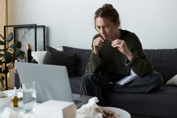 A young woman sits on her couch, lost in thought while staring at a laptop screen, feeling isolated.