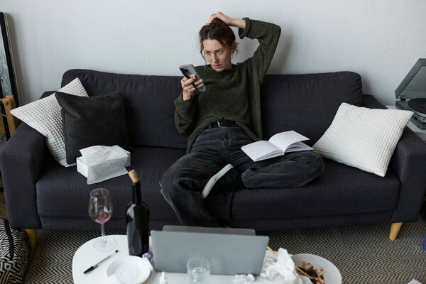 A young woman appears withdrawn on her couch, scrolling through her phone in a quiet home setting.