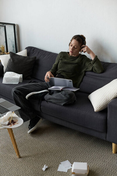 Молодая женщина сидит на своем диване, как потерянный в мыслях среди загроможденного жилого пространства.