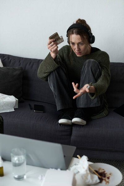 A young woman sits on a couch surrounded by clutter, appearing lost in thought and emotion.