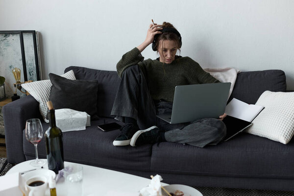 A young woman sits on the couch, feeling overwhelmed as she stares at her laptop, deep in thought.