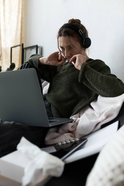 A young woman sits at home, lost in thought with headphones on, staring at her laptop.