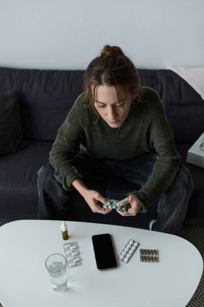 A young woman sits at a table in her living room, holding medication as she deals with her emotions.
