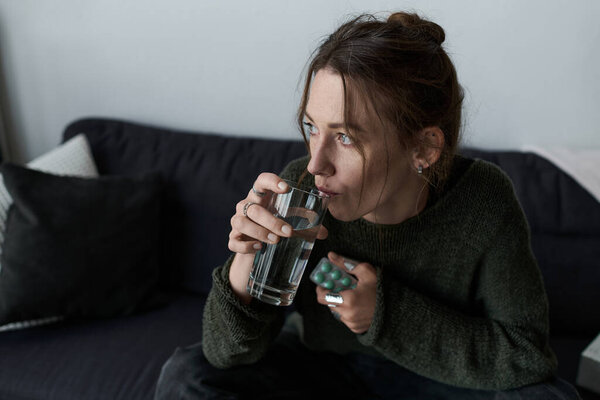 A young woman sits on a couch at home, feeling overwhelmed as she drinks a glass of water.