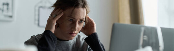A young woman at home looks distressed, holding her head in her hands, staring at her screen.