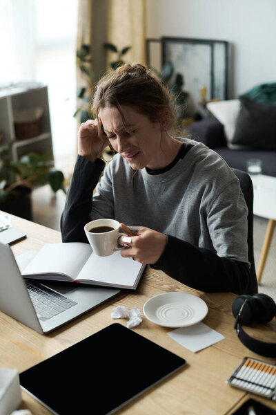 Feeling overwhelmed, a young woman sits at her home desk, holding coffee and looking troubled.
