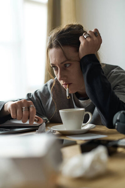 A woman sits alone at a table, visibly distressed while holding a cigarette and staring blankly.