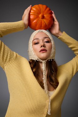 Genç bir kadın kafasında balkabağı ile poz veriyor, şık bir süveter ve şapka giyiyor..