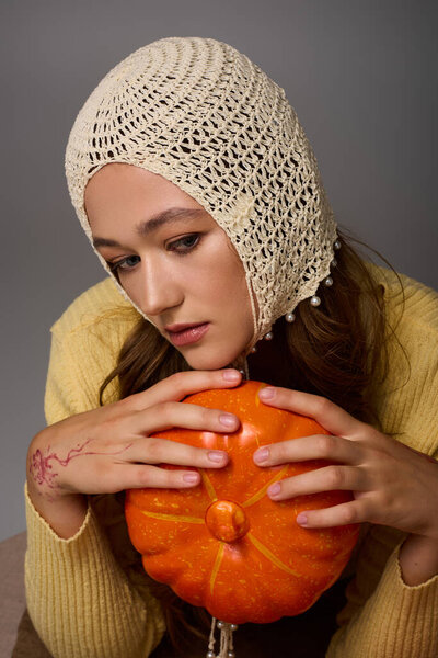 In a stylish outfit, the beautiful young woman holds a pumpkin while posing thoughtfully.