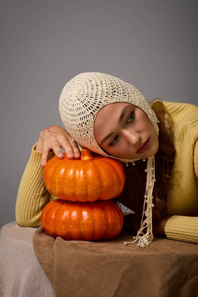 Stylish young woman leans on pumpkins with a thoughtful expression in a warm, inviting atmosphere.