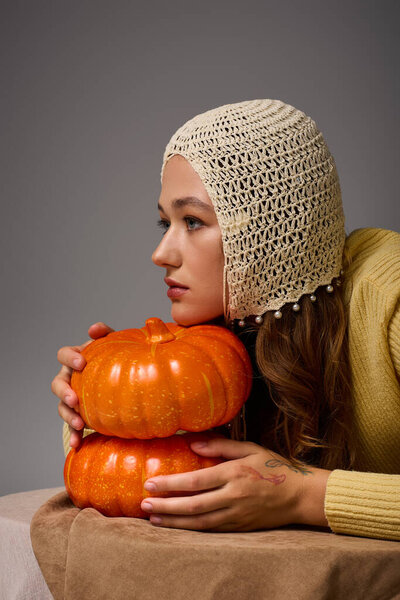 A stylish young woman rests her chin on orange pumpkins, donning a chic hat for a cozy feel.