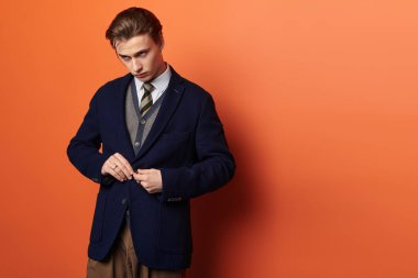 Havalı görünümlü genç bir adam kalın turuncu bir duvarın önünde ceketini ayarlıyor..