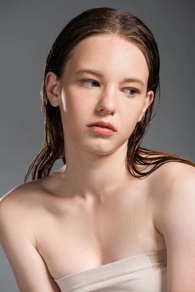 Mujer joven y bonita con la piel mojada y los hombros desnudos mirando hacia otro lado aislado en gris - foto de stock